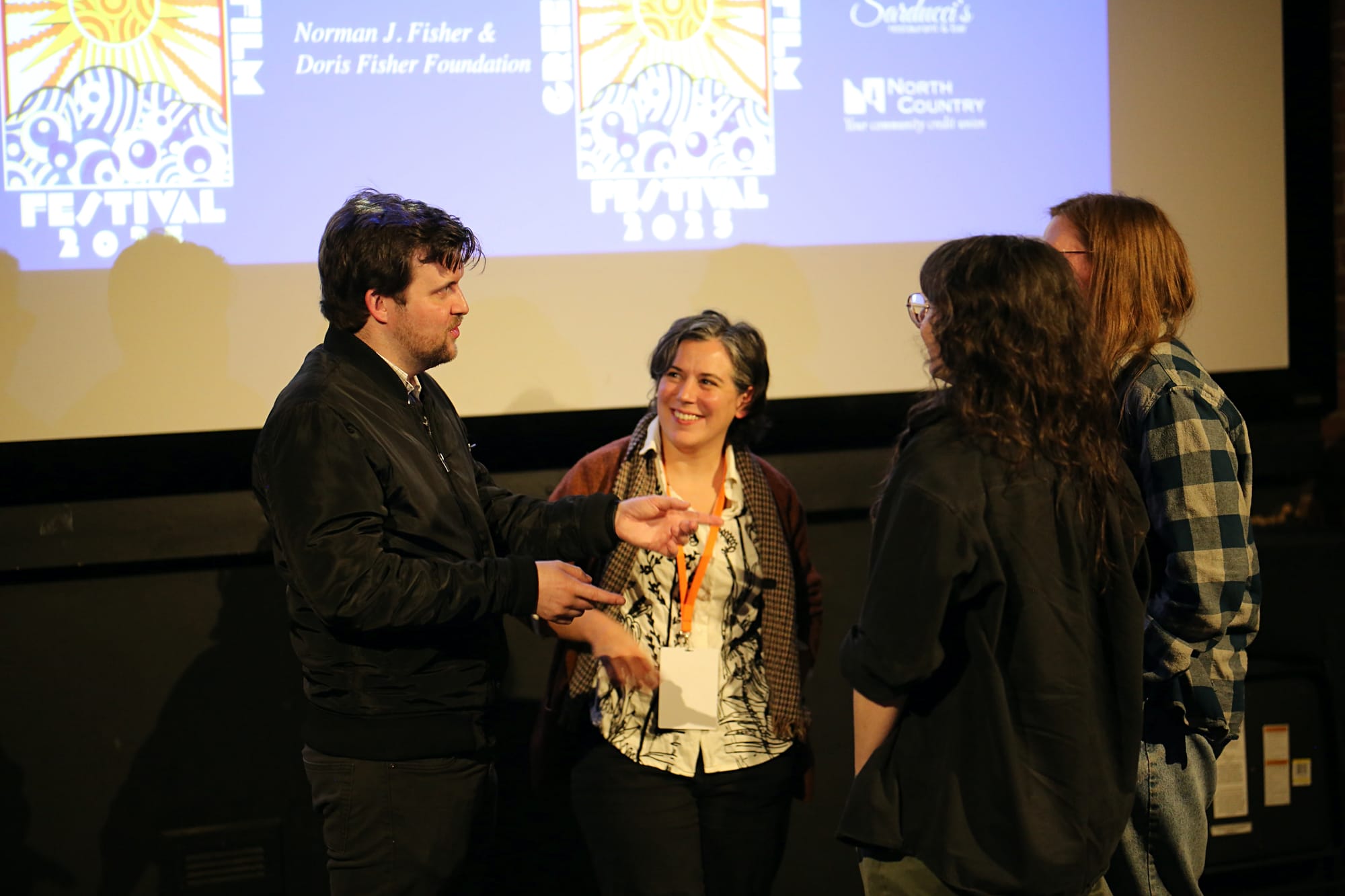 Two guests facing camera chat with a couple not facing the camera, the GMFF logo is projected onto the screen behind them.