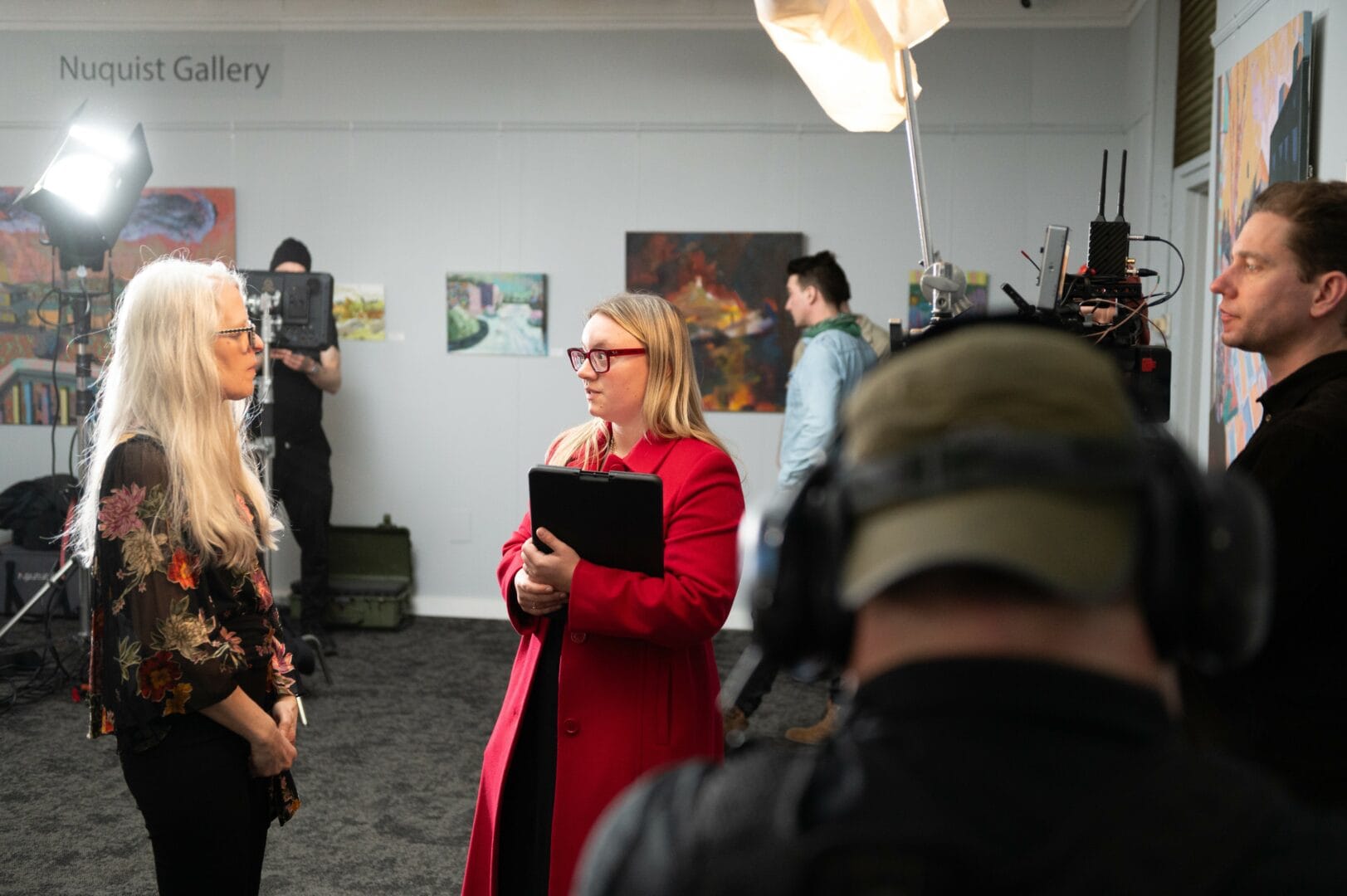 Two women face each other talking as men adjust monitors and lights around them. Paintings are hung on a gray wall behind them.