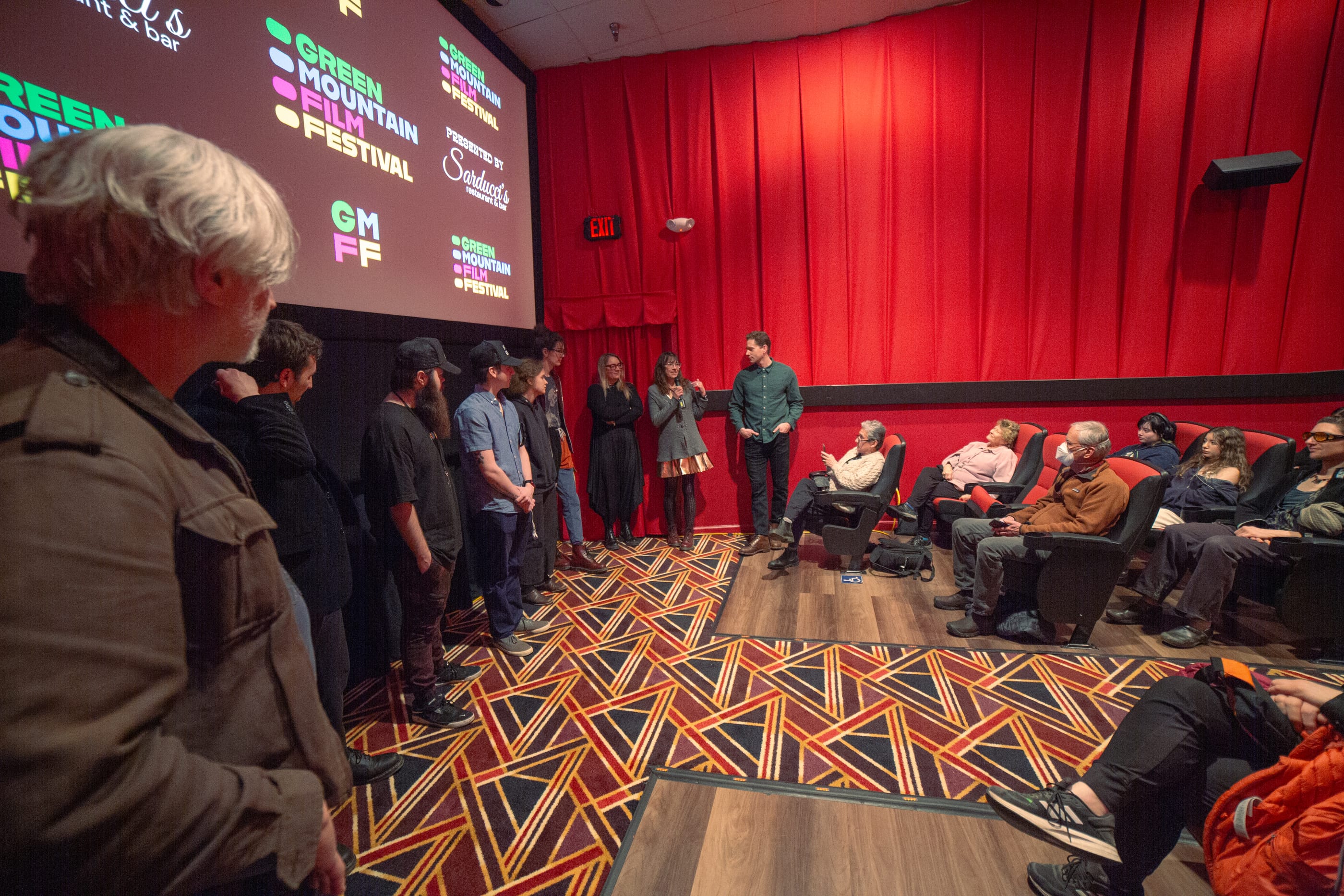 On the left, people are lined up under a screen with the GMFF logo. Right, audience is seated facing them. The back wall is red and the carpet is a multi0colored pattern.